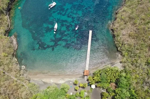 Vista aerea di un lungo molo che si protende in una baia turchese, con diverse imbarcazioni ancorate vicino a una costa boscosa e una piccola spiaggia.