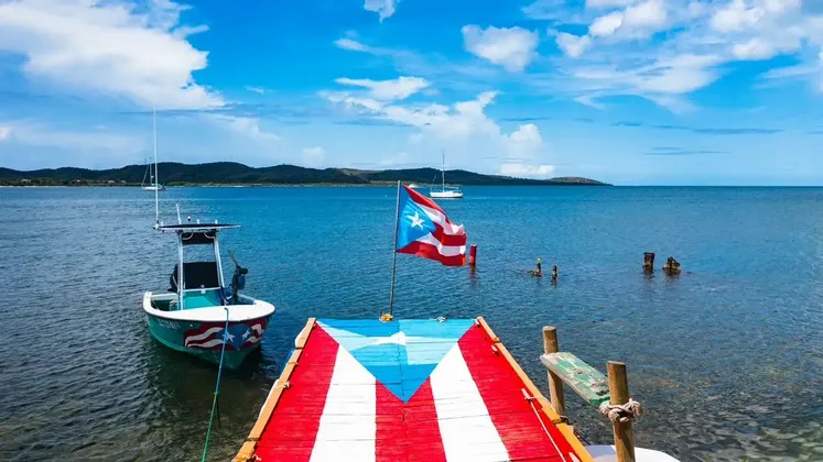 A wooden pier painted as the Puerto Rican flag extends into the sea, with a waving flag and a small boat moored next to it.