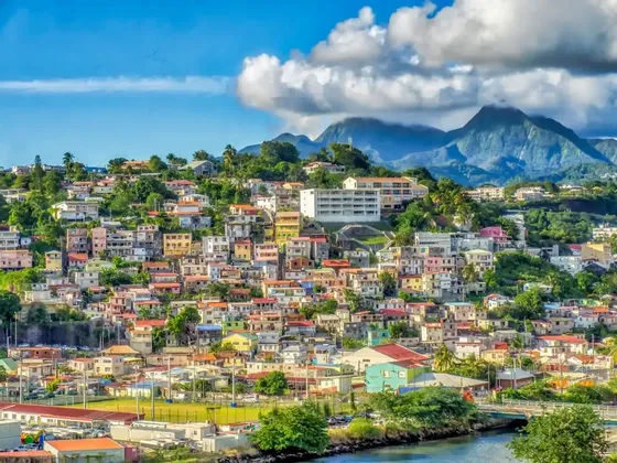 Maisons et bâtiments colorés regroupés sur une colline verdoyante, avec des montagnes luxuriantes en arrière-plan sous un ciel partiellement nuageux.