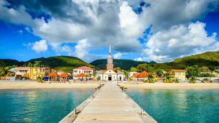 Un quai en bois s'étend sur une eau turquoise vers un village côtier avec une église sur une plage de sable, adossé à des collines verdoyantes.