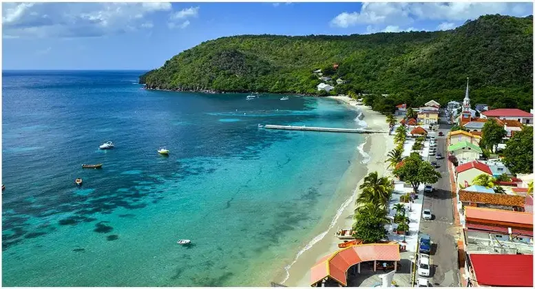 Vue aérienne d'une ville côtière avec une plage de sable, une jetée s'avançant dans une eau turquoise et une colline verdoyante.