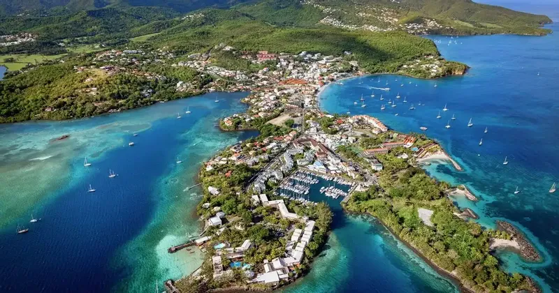 Vue aérienne d'une ville côtière sur une presqu'île, dotée d'un port de plaisance rempli de bateaux, entourée d'eaux turquoise et de collines verdoyantes.