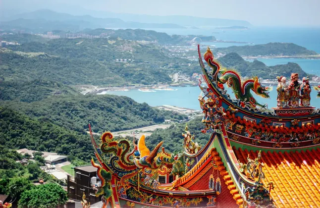 Ein reich verziertes, farbenfrohes Dach eines traditionellen asiatischen Tempels überblickt eine Bucht, grüne Hügel und eine entfernte Küstenstadt.