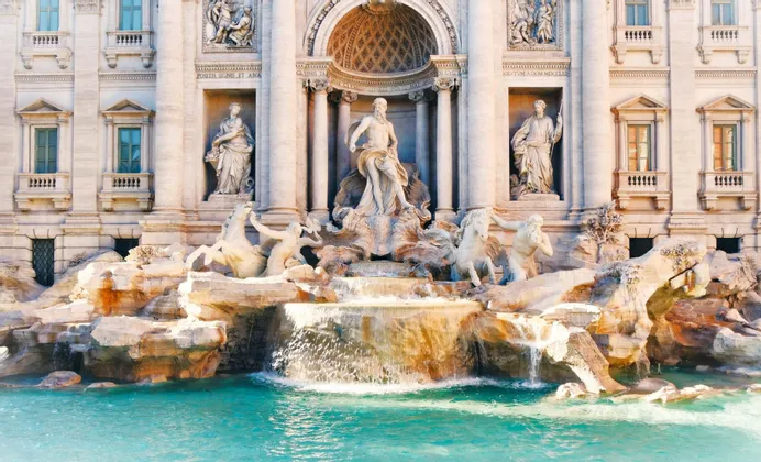Ein prächtiger weißer Marmorbrunnen mit aufwendigen Skulpturen von Figuren und Pferden, dessen Wasser in ein türkisfarbenes Becken fließt.