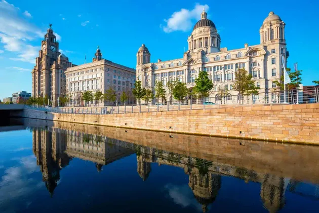 Palazzi storici riccamente decorati, inclusa una torre dell'orologio, che si riflettono nell'acqua placida di un canale sotto un limpido cielo azzurro.