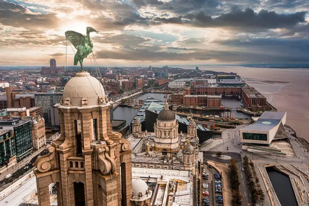 Una vista aerea di una grande statua di uccello in bronzo su una torre, che si affaccia su una storica città portuale e un fiume al tramonto.