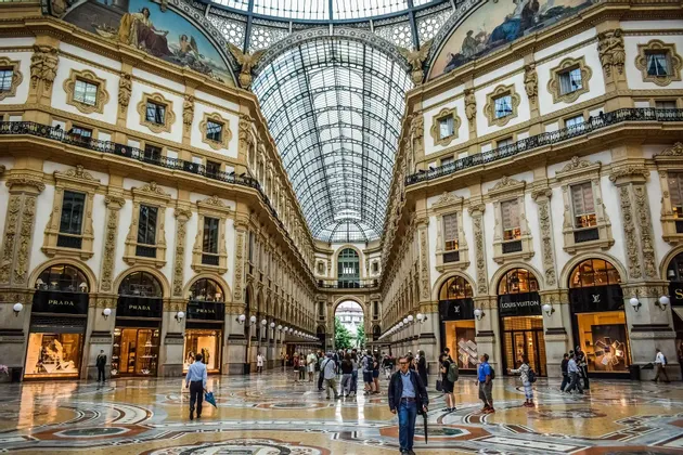 La gente camina por una gran galería comercial con un techo de cristal arqueado, pisos de mosaico y detalles arquitectónicos ornamentados.