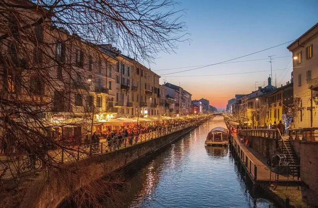 Un largo canal de ciudad al atardecer, con restaurantes iluminados y gente paseando a lo largo de la orilla del agua bajo un cielo colorido.