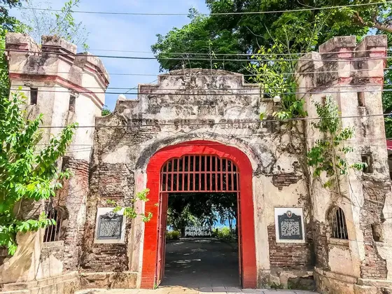 Un portone di fortezza in pietra logorata, con un ingresso ad arco rosso vivo e aperto, invaso da piante verdi.