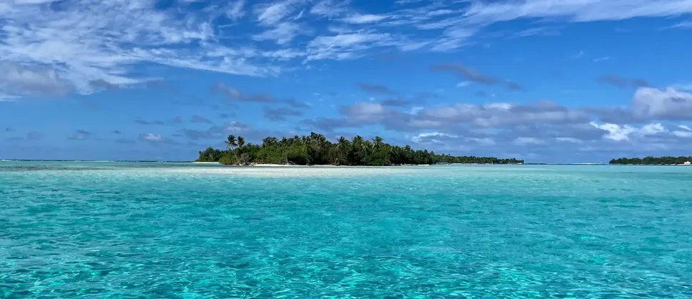 Una piccola isola tropicale coperta di palme è circondata da acque turchesi limpide e poco profonde sotto un cielo blu con nuvole bianche.