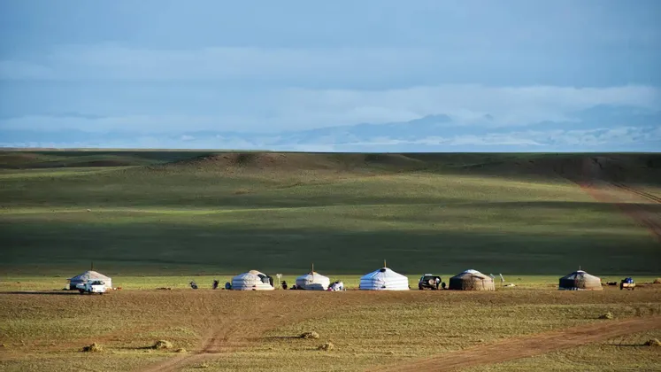 Una fila di yurte tradizionali e veicoli su una vasta steppa erbosa con colline ondulate e montagne in lontananza, sotto un cielo blu.