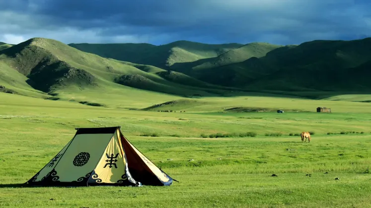 Una tenda a fantasia è montata in un vasto prato verde, con un cavallo al pascolo e dolci colline sotto un cielo drammatico e cupo.