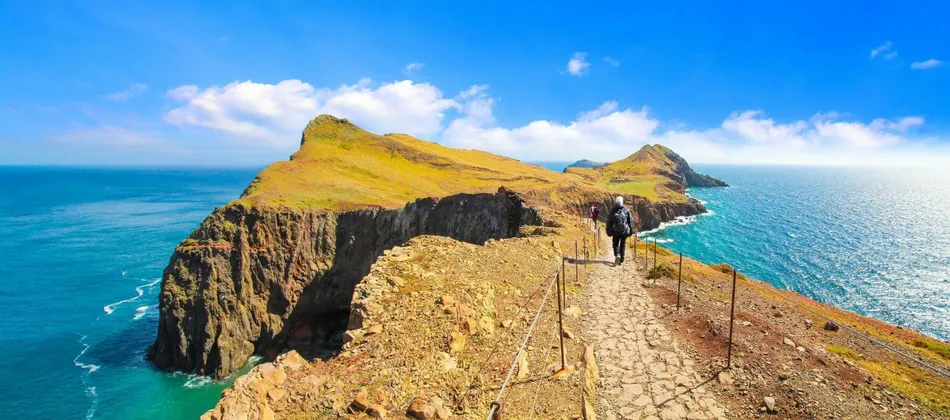 Ein Wanderer mit Rucksack wandert auf einem schmalen Steinpfad entlang einer hohen, grasbewachsenen Küstenklippe mit Blick auf das Meer.