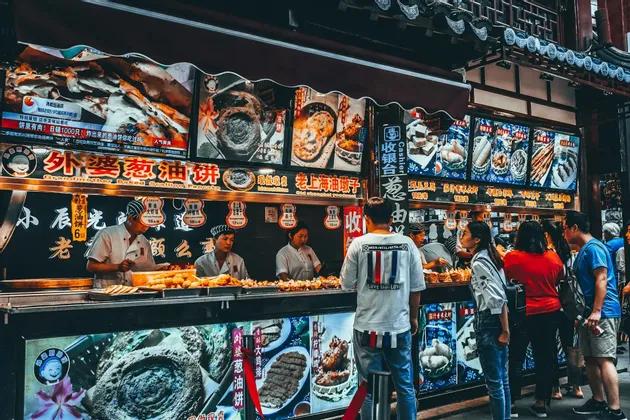 Una folla di persone fa la fila a una bancarella di street food vivacemente illuminata dove i cuochi preparano varie pietanze.