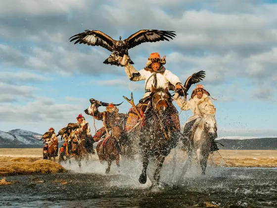 Un gruppo di cacciatori di aquile, vestiti con abiti tradizionali di pelliccia, cavalca i propri cavalli attraverso l'acqua che schizza, ciascuno con un'aquila reale.