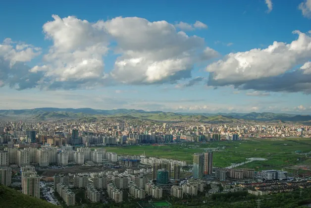 Una vasta città in una valle verde con dolci colline in lontananza, vista dall'alto sotto un cielo parzialmente nuvoloso.