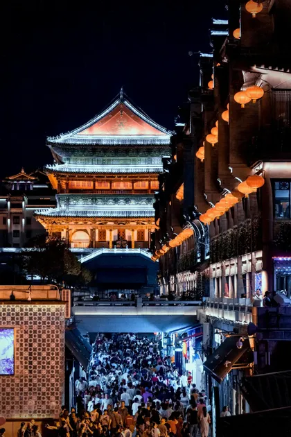 Un grande edificio tradizionale asiatico illuminato si affaccia su una strada cittadina affollata di persone sotto un cielo notturno buio.