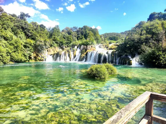 Una cascada de varios niveles cae en un río turquesa claro rodeado de exuberantes árboles verdes bajo un cielo azul soleado.