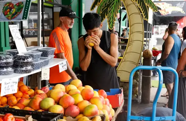 Un uomo in canotta nera annusa un mango giallo a una bancarella di frutta in un mercato all'aperto.