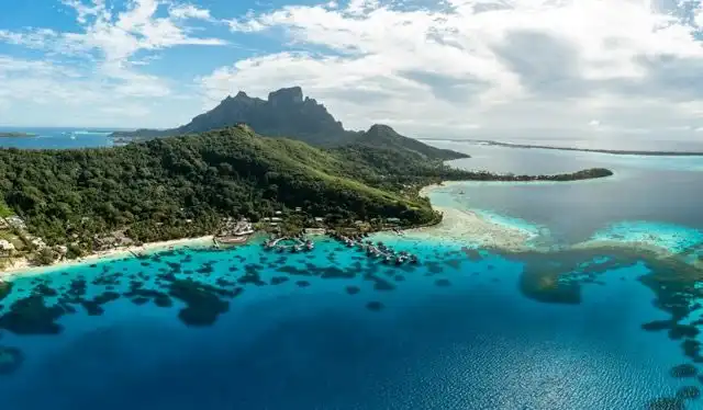 Vista aerea di un'isola tropicale e montuosa con bungalow sull'acqua e una barriera corallina in acque turchesi.