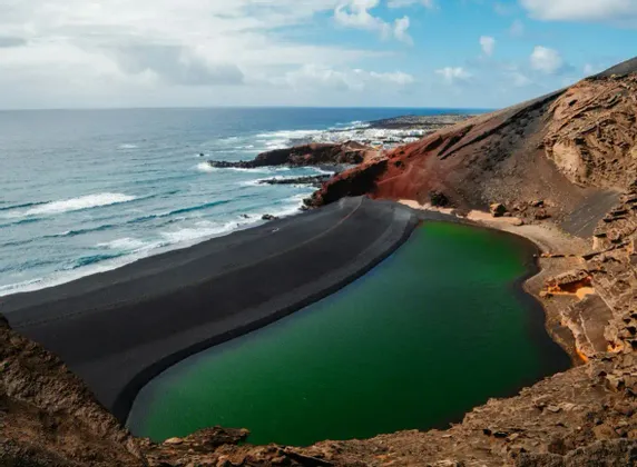 Una laguna verde separata dall'oceano da una spiaggia di sabbia nera ai piedi di una collina vulcanica.