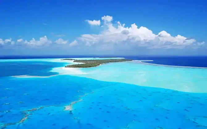 Veduta aerea di un atollo tropicale circondato da una laguna turchese poco profonda e dall'oceano blu intenso, sotto un cielo azzurro con nuvole bianche.