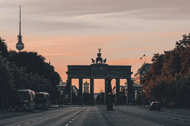 Un arco de triunfo histórico se alza al final de una calle ancha y vacía al atardecer, con una torre de televisión visible al fondo.