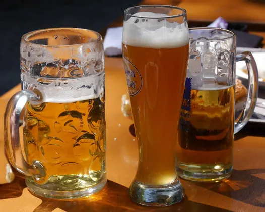 Tres vasos de cerveza diferentes, incluyendo una jarra grande y un vaso alto de cerveza de trigo, están sobre una mesa de madera bajo la luz del sol.
