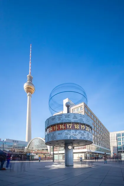Un gran reloj mundial en una plaza de la ciudad, con una alta torre de televisión al fondo bajo un cielo azul claro.
