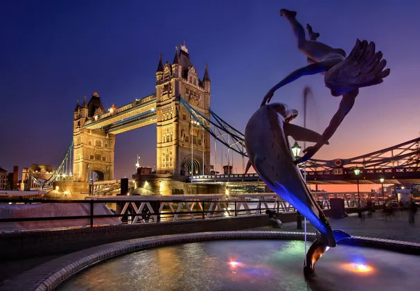 Una escultura de un delfín en una fuente de colores con un gran puente colgante iluminado al fondo durante el crepúsculo.