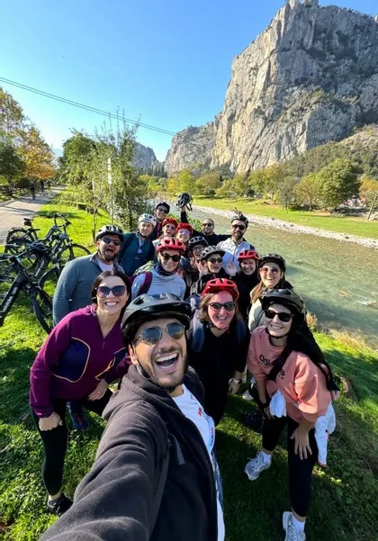 Un gruppo WeRoad in viaggio, con caschi da ciclista, si scatta un selfie su una sponda erbosa del fiume, con ripide montagne sullo sfondo.