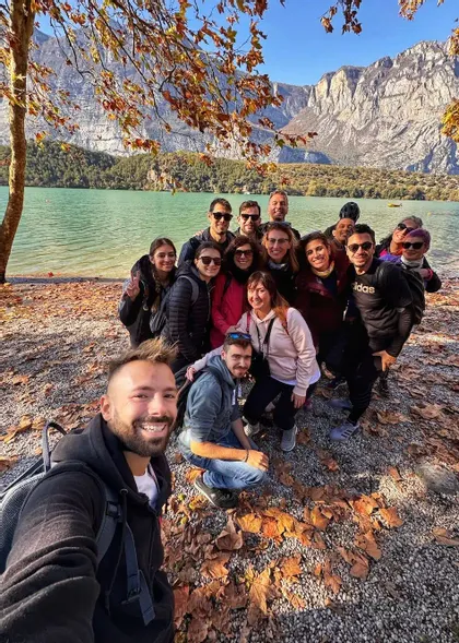Un gruppo WeRoad sorride per un selfie in una giornata autunnale in riva a un lago con montagne sullo sfondo.