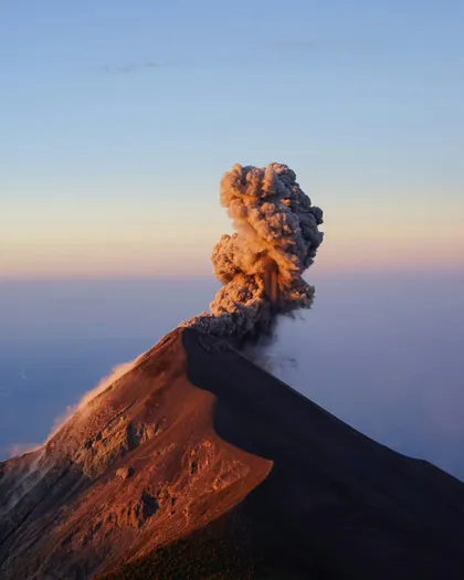 Un vulcano in eruzione invia un'imponente colonna di fumo e cenere nell'aria, illuminata dalla calda luce dell'alba.