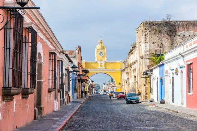 Una strada acciottolata fiancheggiata da colorati edifici in stile coloniale, che conduce verso un grande arco giallo con una torre dell'orologio sotto un cielo sereno.