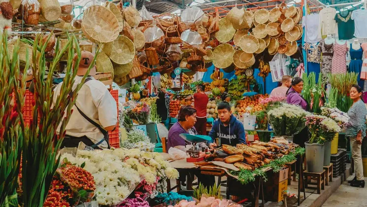 Un vivace mercato coperto dove le persone curiosano tra bancarelle piene di fiori freschi, frutta e cesti intrecciati appesi.