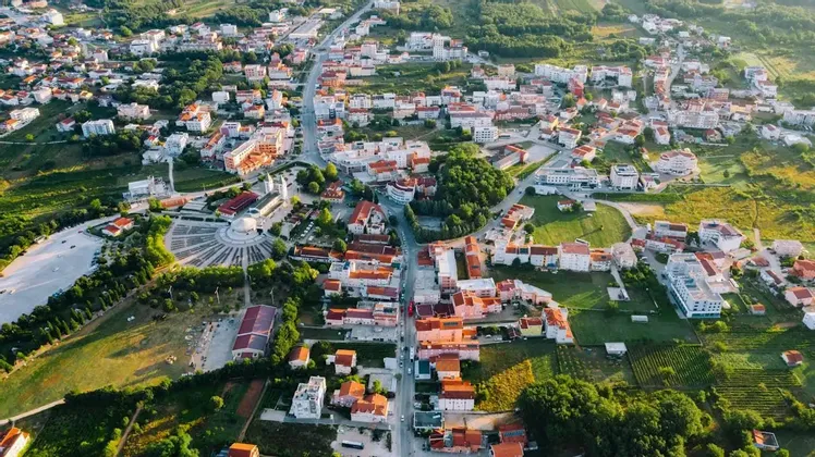 Una vista aerea di una città estesa con tetti di tegole rosse e un'ampia piazza circolare, circondata da rigogliose colline verdi.