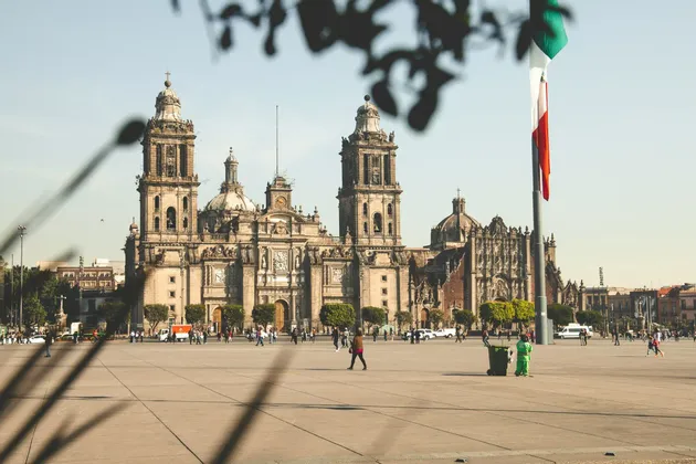 Una grande e sontuosa cattedrale con due campanili si affaccia su un'ampia piazza pubblica soleggiata, con gente che passeggia e una bandiera nazionale che sventola.