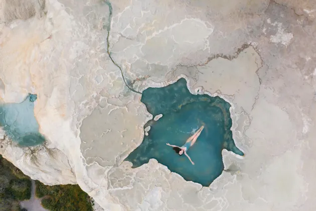 Dall'alto, una donna in costume da bagno blu galleggia sulla schiena in una piscina naturale turchese circondata da terrazze minerali bianche.