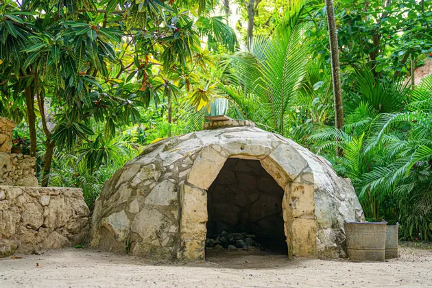 Una struttura in pietra a forma di cupola con un ingresso ad arco si trova su un terreno sabbioso, circondata da una lussureggiante vegetazione tropicale.