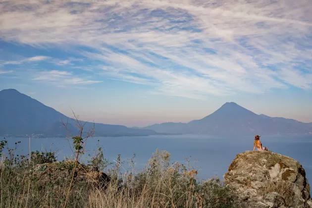 Una persona con un cappello è seduta su una grande roccia, ammirando un vasto lago circondato da montagne sotto un cielo nuvoloso.