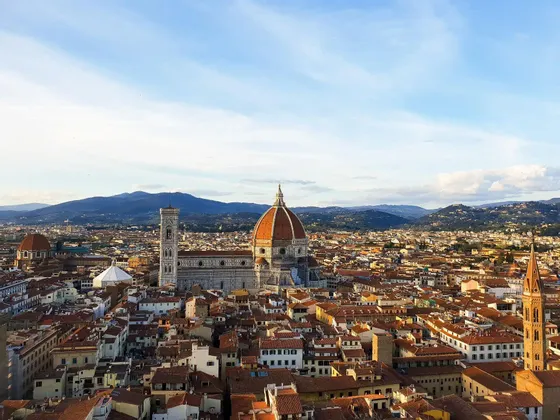 Vista aérea de una ciudad histórica, centrándose en una catedral con cúpula roja y campanario, con montañas visibles en el horizonte.