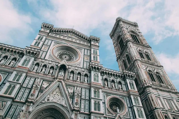 La ornamentada fachada de mármol y el campanario de una catedral, vistos desde abajo contra un cielo azul parcialmente nublado.