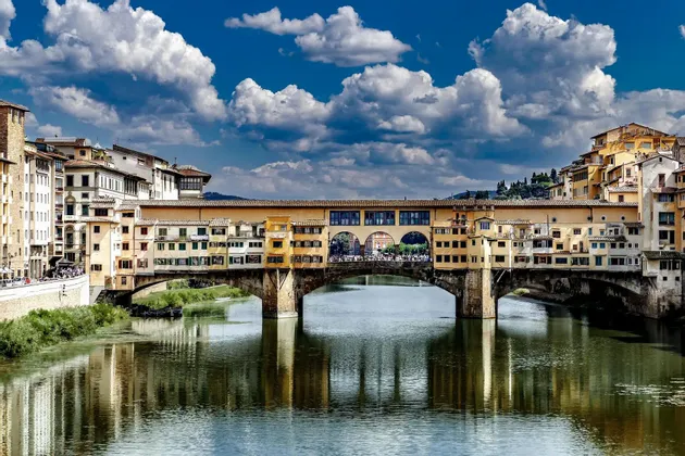 Un histórico puente de piedra, flanqueado por edificios amarillos, cruza un río, con su reflejo visible en el agua bajo un cielo azul y nublado.