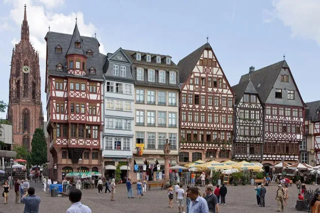 Una plaza histórica europea llena de gente, bordeada de edificios tradicionales de entramado de madera y una torre de catedral gótica al fondo.