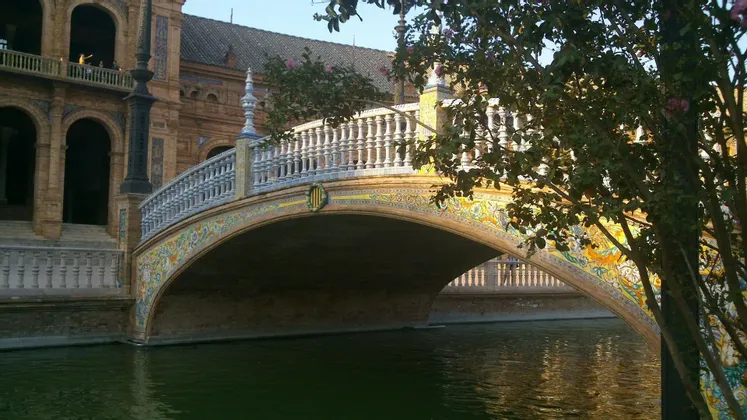 Un ponte ad arco decorato con piastrelle colorate e una balaustra bianca attraversa un canale di fronte a un grande edificio in mattoni.