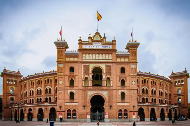 La facciata simmetrica in mattoni rossi dell'edificio della Plaza de Toros, con archi e una bandiera spagnola, vista da una grande piazza sotto un cielo nuvoloso.