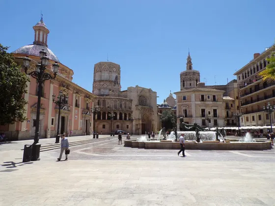 Una piazza cittadina europea illuminata dal sole, con una grande fontana, edifici storici e persone che passeggiano in una giornata serena.