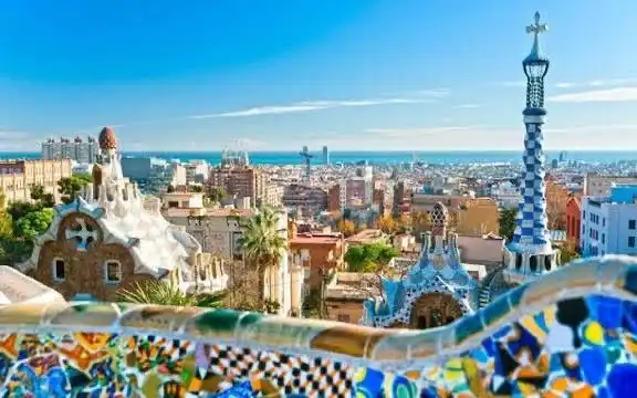 Un paesaggio urbano con edifici unici e bizzarri e una guglia blu e bianca, visto oltre un muro a mosaico colorato.