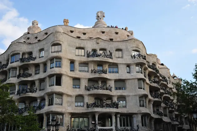 Un edificio in pietra ondulato con balconi in ferro battuto ornati e comignoli scultorei sul tetto, visto contro un cielo azzurro.