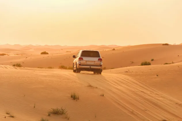 Un SUV argenteo attraversa dune di sabbia ondulate in un vasto paesaggio desertico sotto un cielo caldo e velato al tramonto.